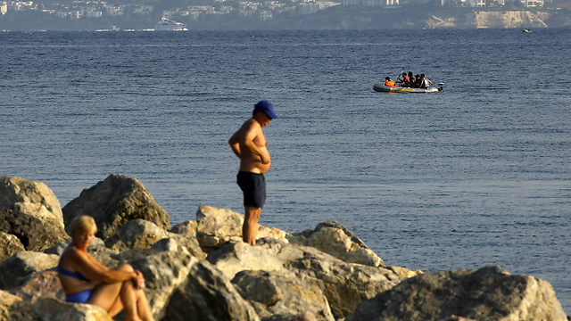 תיירים בקוס על רקע סירת מהגרים שמגיעה לאי (צילום: רויטרס) (צילום: רויטרס)
