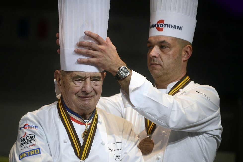 ז'רום בוקוז מסדר את הכובע על ראש אביו, שף פול בוקוז, בגמר של תחרות הקולינריה הבינלאומית ה-14 בשנת 2013, שבה הפרס הראשי מכונה בוקוז ד'אור (  ) (  )