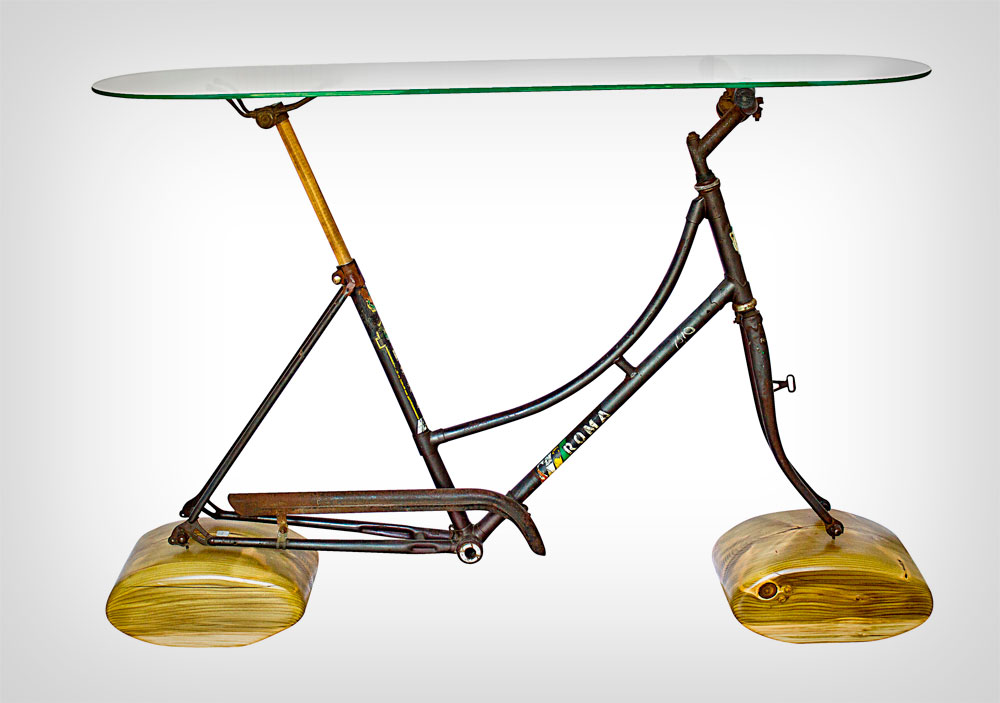 שלדה של אופניים ישנים, ומעליה זכוכית שהופכת את הפריט לשולחן. הרגליים עשויות עץ גושני שרגב מצא בסביבת הסדנה שלו