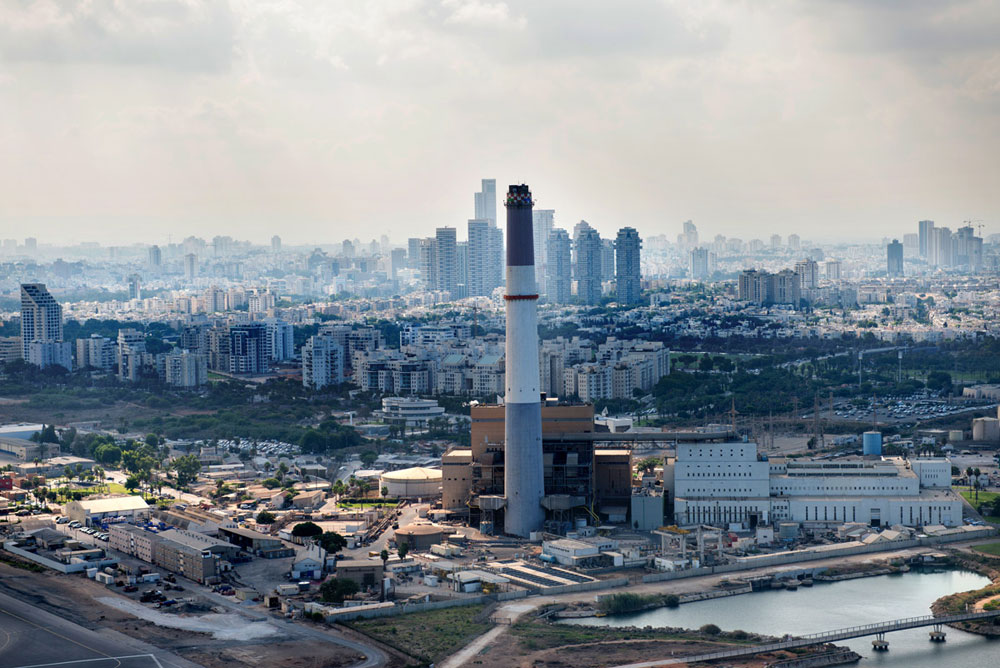 השחר עולה על תחנת הכוח רידינג, שמצולמת כאן מכיוון הים. הארובה לא מעשנת כבר שנים, השטחים סביבה הפכו לטיילת הצפונית של תל אביב, ומאחוריה מגדלי אקירוב ופארק צמרת אילן נחום תל אביב