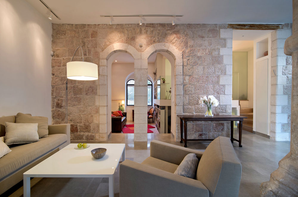 הפתחים המקושתים המקוריים בקיר האבן נפתחו בשיפוץ, לאחר שנאטמו עם השנים. כך נוצר חיבור ויזואלי בין שני חלקי הדירה. בתמונה: מבט מכיוון הסלון לחדר המשפחה והעבודה, שנמצא במורד שלוש מדרגות. מימין עלייה לגלריה שבה חדר השינה של בעלי הבית, עם חדרי רחצה וארונות צמודים. רוזנשיין בחר לשלב ישן בחדש, ולכן נבחרו רהיטים וגופי תאורה מודרניים מתי רוזנשיין דירה שכונת מחנה יהודה